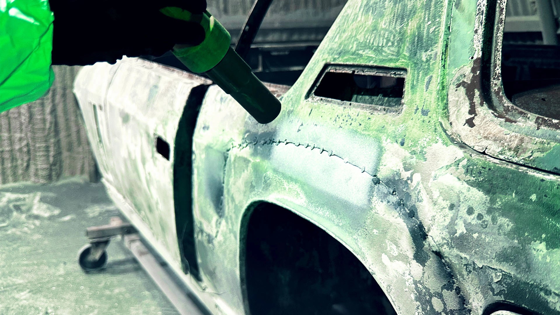 Ein Mitarbeiter der Firma ecoBlast in Nettetal bei der professionellen Entlackung einer Oldtimer-Karosserie mittels Strahltechnik. Das Bild zeigt eine Nahaufnahme der Strahldüse, die präzise alte Farbschichten vom Metall des Fahrzeugs entfernt, während der Anwender grüne Schutzkleidung trägt.
