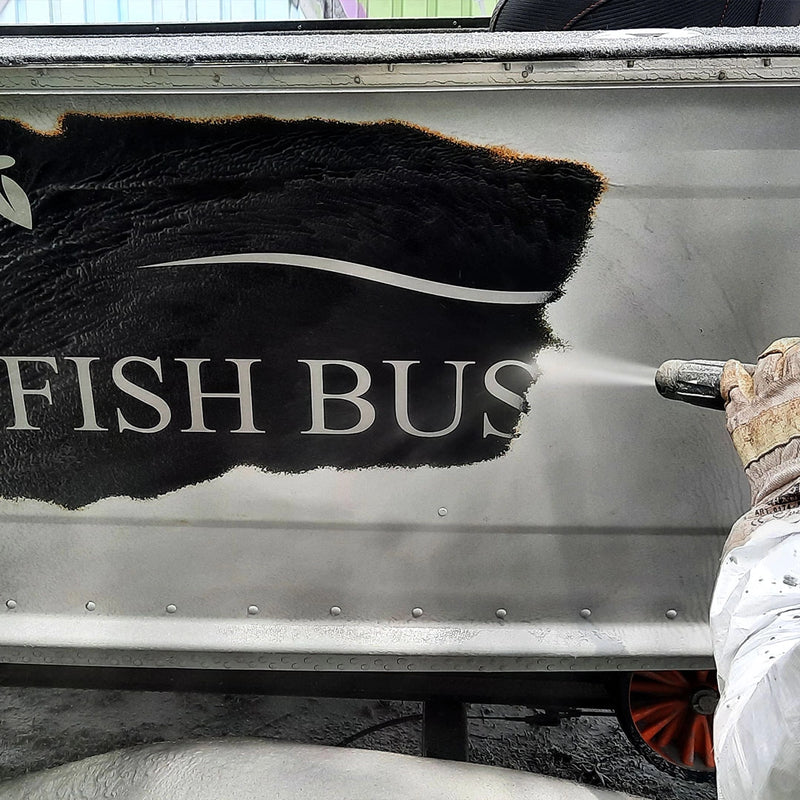 Präzise Oberflächenreinigung durch die Firma ecoBlast in Nettetal. Die Aufnahme zeigt, wie mittels Strahltechnik eine schwarze Farbschicht samt Logo-Schriftzug „FISH BUS“ von der Metalloberfläche eines Fahrzeugs entfernt wird, um den Untergrund schonend freizulegen.