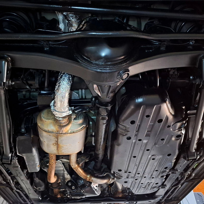 Detailansicht eines frisch versiegelten Fahrzeugunterbodens nach der Behandlung durch die Firma ecoBlast in Nettetal. Das Bild zeigt das sauber beschichtete Differential und angrenzende Fahrwerksteile in Schwarz, während die Abgasanlage und hitzeempfindliche Bereiche zum Schutz während des Versiegelungsprozesses sorgfältig mit Alufolie maskiert wurden.